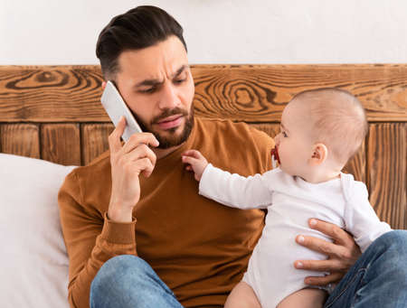 Young Father Talking On Phone Sitting With Baby Toddler In Bedroom At Home. No Personal Life For Dad, Little Child Reaching For Daddys Smartphone. Paternity Leave, Fatherhood Difficulties And Problemの写真素材