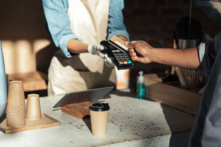 Pay for takeaway coffee at social distancing. African american lady in apron and protective gloves...