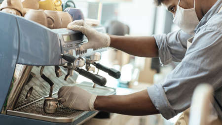 Takeaway coffee in modern cafeteria in morning during COVID-19 pandemic and social distancing. Millennial african american man in apron, gloves and protective mask make cappuccino, cropped, panoramaの写真素材