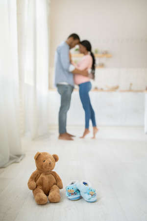 Teddy bear and cute baby booties on floor indoors and black pregnant couple hugging on background, selective focus. Child toy with tiny shoes and young expectant family at home, copy spaceの写真素材