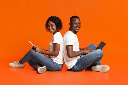 Cheerful black couple sitting back to back on floor and using social media over orange studio background, copy space. African american happy woman with digital tablet and man with laptopの写真素材