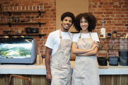 Small family business concept during covid-19 epidemic. Smiling millennial african american man and woman baristas in aprons look at camera in modern loft cafe interior with equipment, copy spaceの写真素材