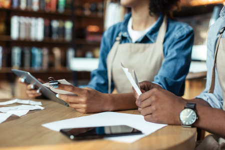 Cafe shop, small business, accounting and bill payment. Focus on hands of african american millennial male and female, owners with tablet and papers in hands on table with smartphone, croppedの写真素材