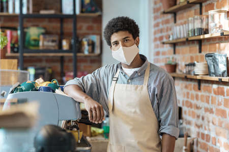 Coffee business, startup and back to normal concept. Young african american guy barista in apron and protective mask with equipment in coffee shop in morning at lockdown, quarantine, coronavirusの写真素材