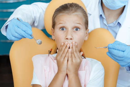 Work with children, dental examination and orthodontics. Scared kid in chair covers mouth with hands and expresses fear from doctor in white coat, protective mask and gloves with medical toolsの写真素材