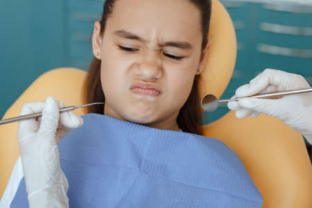 Natural children emotions and toothache treatment. Little hispanic girl in medical chair looks with...