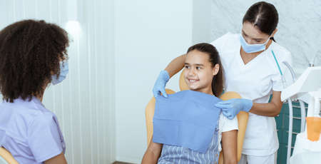 Preparation for examination and treatment of oral cavity, orthodontics and pediatric dentist. Nurse puts napkin on girl, african american doctor talking to little girl in clinic, panorama, copy spaceの写真素材