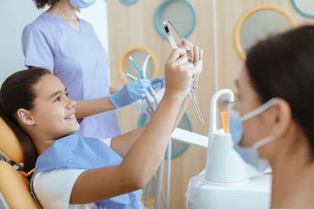 Satisfied dentist clinic patient look at her perfect smile in mirror after treatment in office with medical equipment in background and nurse in white coat, protective mask and rubber gloves, croppedの写真素材