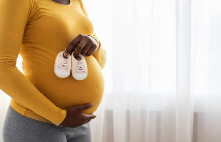 Motherhood concept. Closeup of cute baby shoes on pregnant black lady big belly. Unrecognizable...