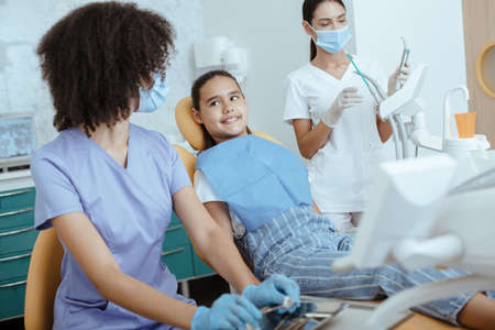 Dentist clinic. Hands of unrecognizable african american pediatric doctor in protective mask and gloves with nurse making examination procedure for smiling cute little girl, sit on chair in hospitalの写真素材
