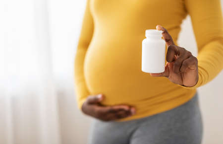 Closeup of black pregnant woman holding jar with pills or vitamins and touching her big tummy. Unrecognizable african american expecting lady recommending supplements for future mothers, copy spaceの写真素材