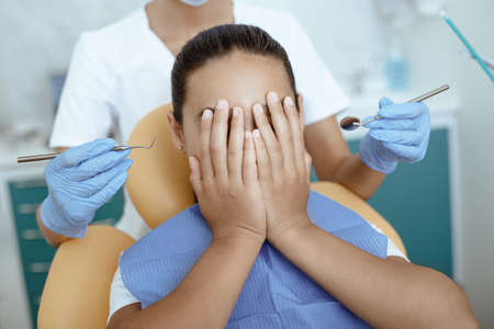 Modern dental treatment and fear. Scared girl covers face with hands and does not allow doctor in...
