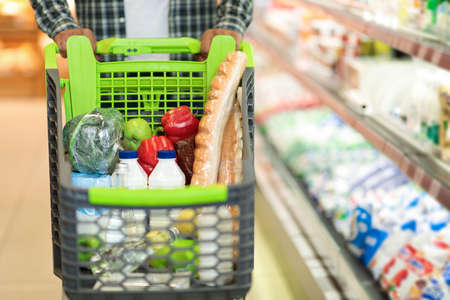 Black Man With Shopping Cart Buying Food Products In Supermarket, Holding Full Trolley With Vegetables, Dairy And Bread. Customer Buys Grocery In Store Concept. Cropped, Selective Focus, Copy Spaceの写真素材