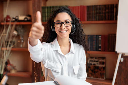 Happy For Success. Smiling woman wearing headphones, showing thumbs up gesture, enjoying e-learning sitting at desk. Excited lady in headset recommending distance education, looking at cameraの写真素材
