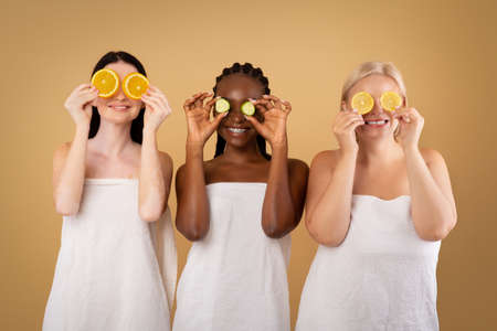 Beauty And Skin Care. Three Diverse Women In Bath Towels Covering Eyes With Cucumber And Orange Slices, Making Organic Face Masks, Posing Together Over Beige Studio Background, Copy Spaceの写真素材