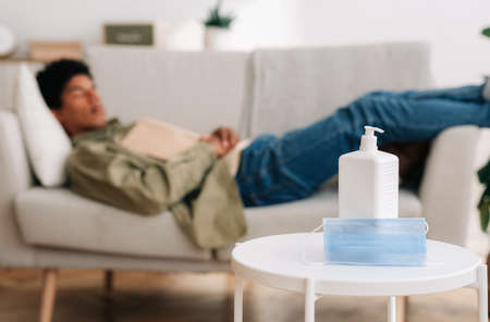 E-learning during covid-19 lockdown concept. Blurred black teenager sleeping on sofa with textbook, selective focus of medical mask and antiseptic on table. Blank spaceの写真素材