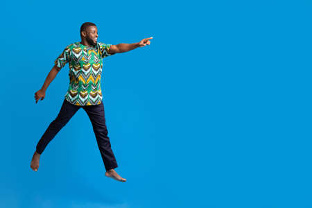 Excited black man in colorful authentic costume running towards copy space for advertisement, blue...