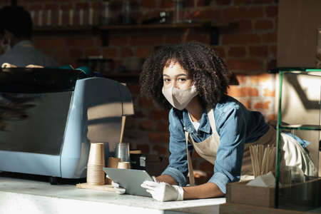 No customers in modern cafe during covid-19 lockdown. Bored millennial african american lady in apron and protective mask and gloves with tablet in hands on bar with equipment in morning, copy spaceの写真素材