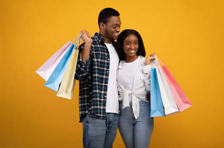 Discounts, sales and modern shopping at leisure time. Cute millennial african american man and woman hug and hold colorful purchases bags, isolated on yellow background, studio shot, copy spaceの写真素材