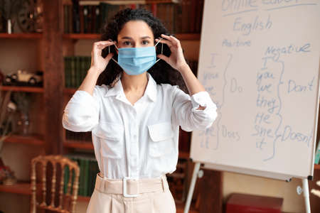 Education And Protection Concept. Female teacher wearing medical surgical mask to prevent infection, illness, flu or covid-19, putting it on face at classroom. Pandemic, Health Care And Quarantineの写真素材
