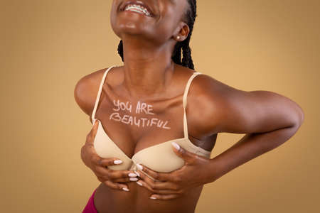 Love Your Body. Happy Black Woman In Bra With You Are Beautiful Inscription On Chest Standing Over Beige Studio Background, Smiling With Dental Braces And Enjoying Her Beauty, Cropped Imageの写真素材