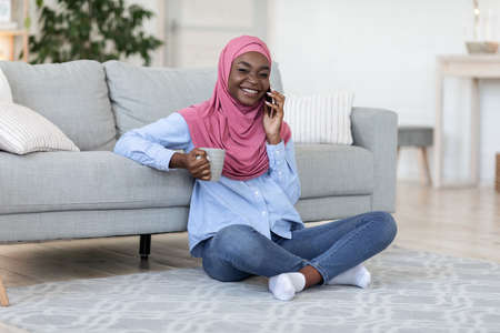 Domestic Pastime. Young Black Muslim Woman In Hijab Enjoying Tea And Talking On Phone At Home,...