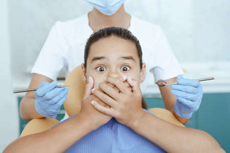 Emotions and fear of dental treatment, examination of teeth and put filling. Scared kid in chair with wide open eyes covers mouth with hands and female doctor in coat and gloves, close up, croppedの写真素材