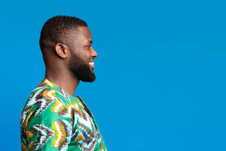 Side view of black smiling guy in african shirt looking at free space on blue, studio shot,...