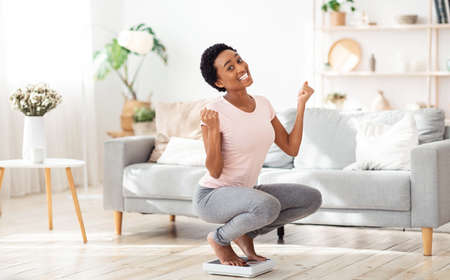 Excited black woman sitting on scales at home, happy with result of her slimming diet, copy space. Emotional African American lady achieving her weight loss goal, gesturing YESの写真素材
