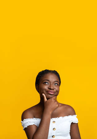 Portrait Of Pensive African American Lady Looking Up At Copy Space On Yellow Background, Curious Black Woman Touching Chin, Thinking About Interesting Offer And Playfully Smiling, Vertical Shotの写真素材