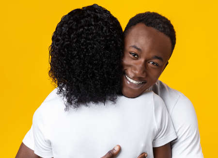 Happy black man hugging his girlfriend and smiling at camera over yellow studio background, closeup. Thankful african american young man embracing his woman. Love and affectionate conceptの写真素材