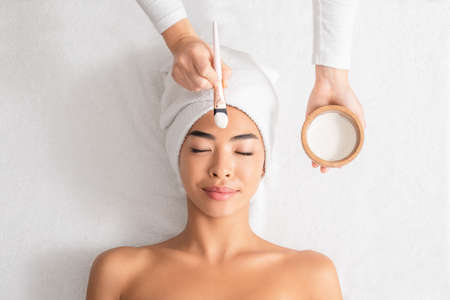 Beauty Treatments. Cosmetologist applying clay mask on asian woman face at spa salon, relaxed korean lady with towel on head enjoying wellness day at beauty center, top view with free spaceの写真素材