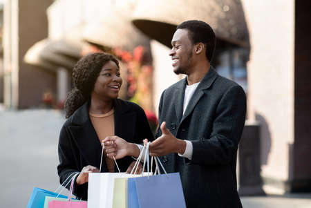 Love, holiday sales, shop, retail, consumer. Cheerful millennial african american man and lady with many coloured shopping packages, guy shows things in bag after shopping in city mall, free spaceの写真素材