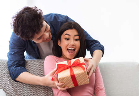 Asian Husband Giving Wrapped Christmas Gift To Wife Surprising Her With Xmas Present Sitting On Sofa...