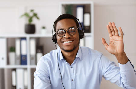 Office worker concept. Happy african american guy consultant with headset waving at camera, office...
