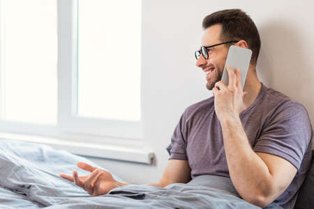Happy Man Talking On Mobile Phone Calling Somebody Having Cellphone Conversation Sitting In Bed In Bedroom At Home. Always In Touch, Guy Speaking On Smartphone. Copy Spaceの写真素材