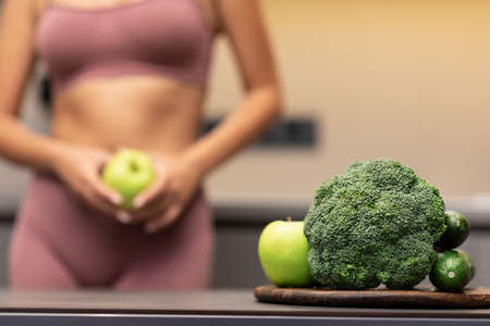 Weight Loss And Dieting. Skinny Girl Dieting Holding Apple Standing In Kitchen, Focus On Green Vegetables On Table. Weight-Loss Diet, Calories, Healthy Detox Food Recipes Concept. Croppedの写真素材