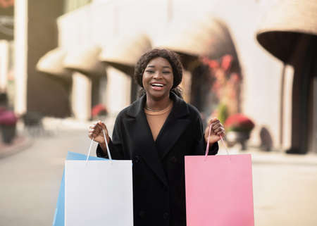 Shopaholic and huge sale at Black Friday. Smiling millennial african american woman in coat with colored paper shopping bags with purchases and enjoys new clothes, near city mall, copy spaceの写真素材