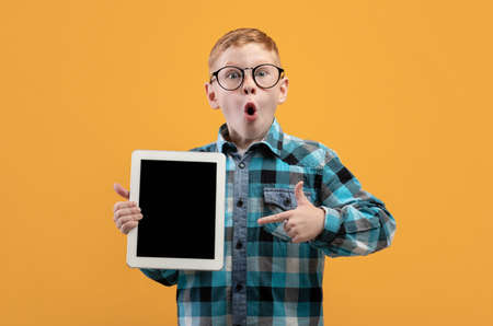 Amazed schoolboy in glasses pointing at empty digital tablet screen, recommending cool educational...