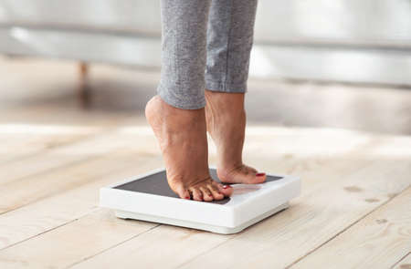 Cropped view of young black woman standing on scales at home, measuring her weight, closeup of feet....
