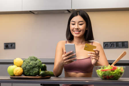 Grocery Shopping App. Asian Woman With Phone And Credit Card Buying Groceries Food Online Using Smartphone Application Sitting In Modern Kitchen At Home. Lady Ordering Food, Delivery Service Conceptの写真素材