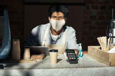 Contactless payment, good service in cafe during social distance and COVID-19. Millennial african american male waiter in apron, protective mask and gloves giving terminal and cup of coffee to clientの写真素材