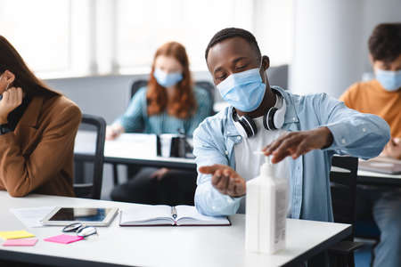 Hygiene And Protection Concept. Black student guy applying antibacterial sanitizer on hands from bottle, wearing surgical face mask, sitting in classroom. Prevent the spread of infection and germsの写真素材