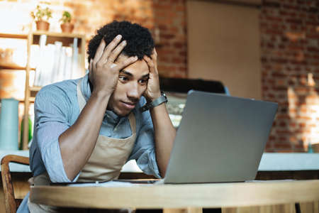 Ruin and financial problems during covid-19 quarantine. Sad young african american guy in apron holds head and looks in laptop at financial accounts and big debt in modern cafe, empty spaceの写真素材