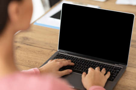 Lifestyle And Gadgets Concept. Over the shoulder view of woman using laptop with blank black screen for mockup template, sitting at wood table, back shot. Person typing on computer keyboardの写真素材