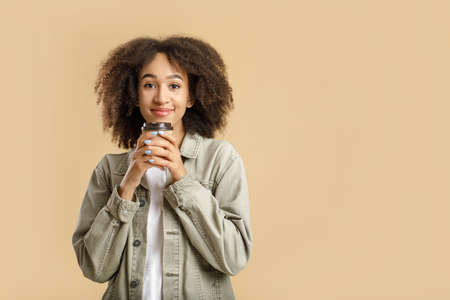 Favorite hot drink and break. Happy millennial curly african american lady in casual, inhaling scent of coffee in takeaway paper cup, isolated on yellow background, free space, studio shotの写真素材