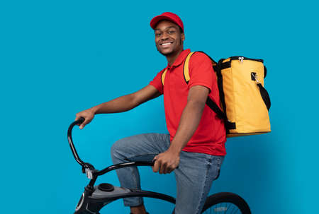 Fast Delivery Service. Smiling african american courier guy in red cap and uniform riding bicycle and wearing yellow thermo backpack bag isolated on blue studio background. Shipment and postの写真素材