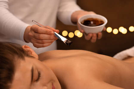 Unrecognizable massage therapist applying exfoliating body mask with brush on relaxed woman back at spa, body care concept. Smiling young lady having body treatment with aroma at modern spa, closeupの写真素材