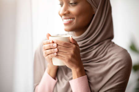 Cropped view of happy black woman in hijab warming her hands on cup of hot coffee near window at home. Lovely young Muslim lady having relaxing morning with aromatic drink, enjoying her breakの写真素材