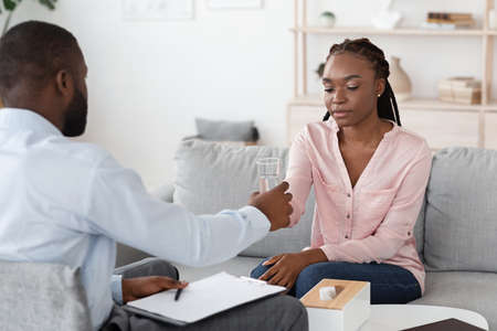 Psychotherapist Comforting Upset African American Lady During Therapy Session, Giving Glass Of Water, Young Black Woman With Mental Problems Sitting On Couch At Counselors Office, Free Spaceの写真素材